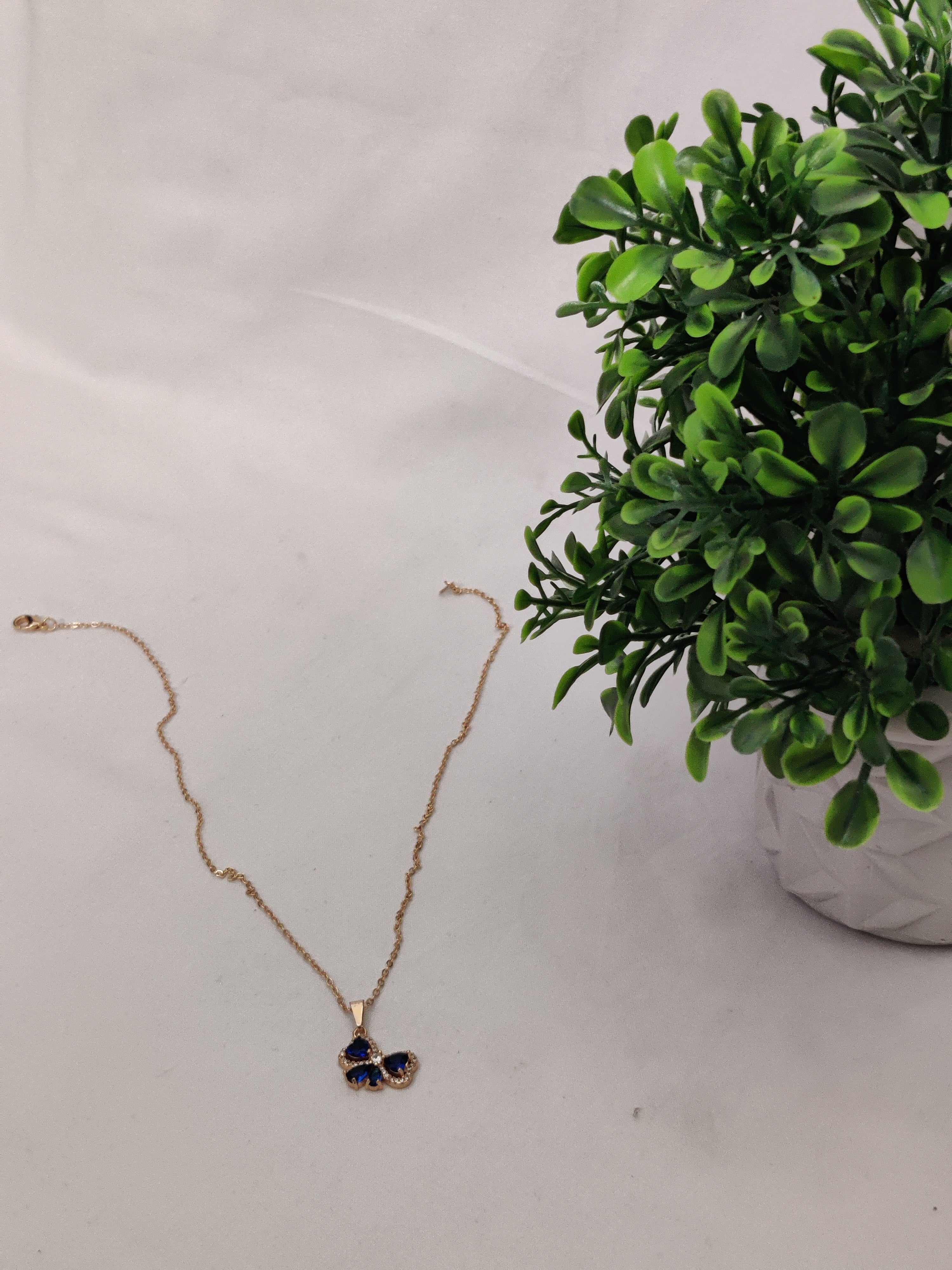 Gold chain necklace with a blue butterfly gemstone pendant placed on a white background next to green leaves